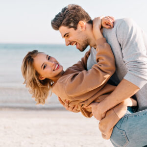 Short-haired blonde lady embracing husband on sea background. Outdoor photo of good-humoured man dancing with girlfriend near ocean.